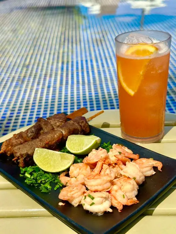 Espetinhos de carne, camarões e limão com um drink de laranja à beira da piscina