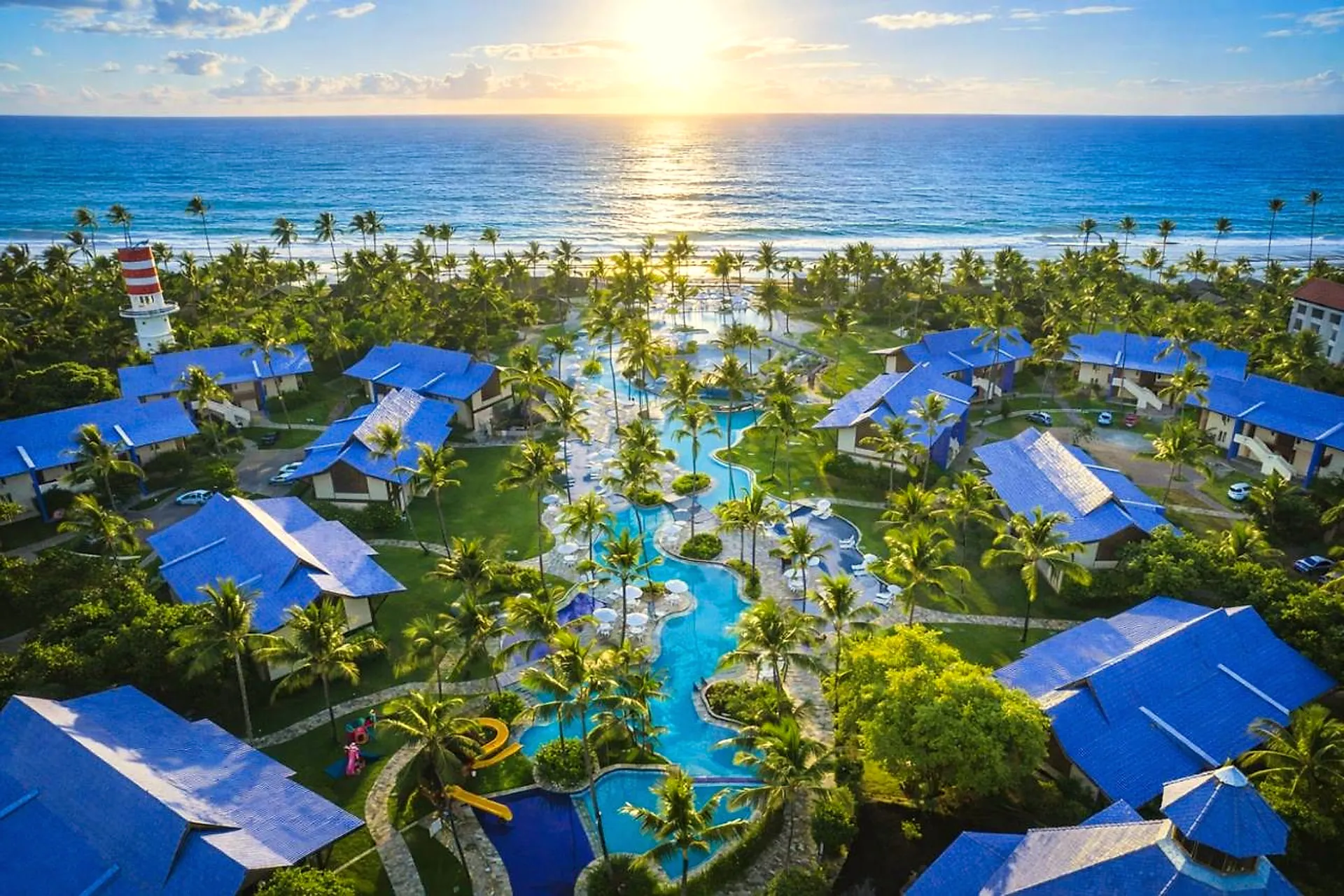 Vista aérea do resort ao amanhecer, com bangalôs de telhado azul, piscinas entre coqueiros e o mar ao fundo