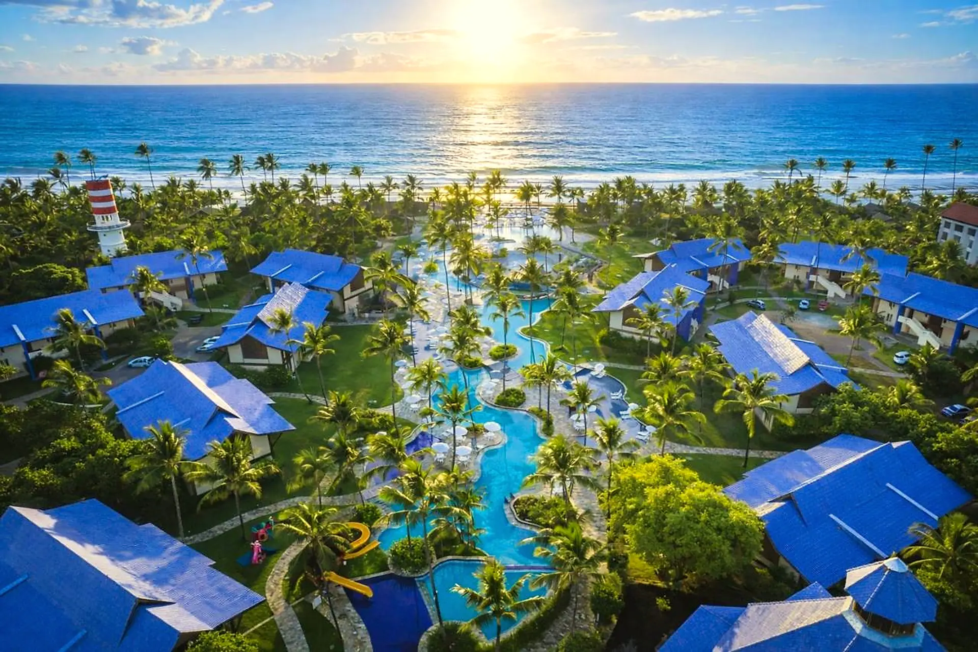 Vista aérea do resort ao amanhecer, com bangalôs de telhado azul, piscinas entre coqueiros e o mar ao fundo